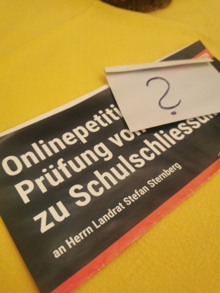 Ein Flyer der Online-Petition liegt mit einem aufgehefteten Fragezeichen auf einem gelben Tischtuch.