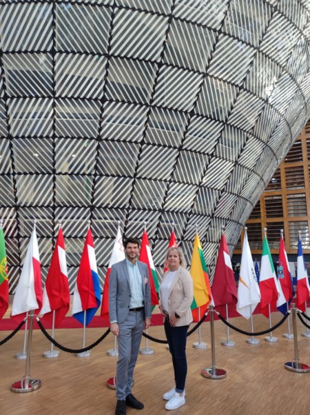 Christine Klingohr und Christian Winter stehen im Foyer des Ratsgebäudes der EU vor den Nationalfahnen der Mitgliedsstaaten.