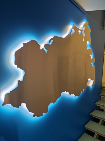 Der Umriss des Landes Mecklenburg-Vorpommerns hebt sich silbern und mit Hintergrundbeleuchtung von der blauen Wand ab.